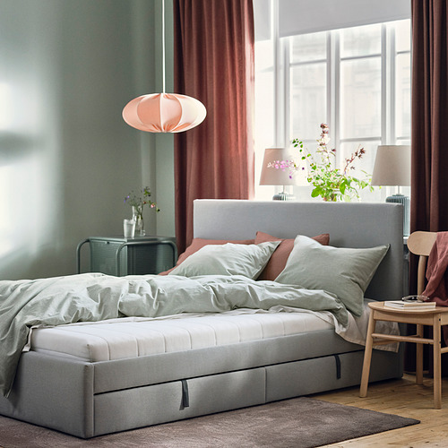 Acogedor dormitorio con colchón ÅFJÄLL gris sobre suelo de madera cerca de la ventana. Iluminación suave de lámpara rosa, paredes verdes y cortinas rosas.