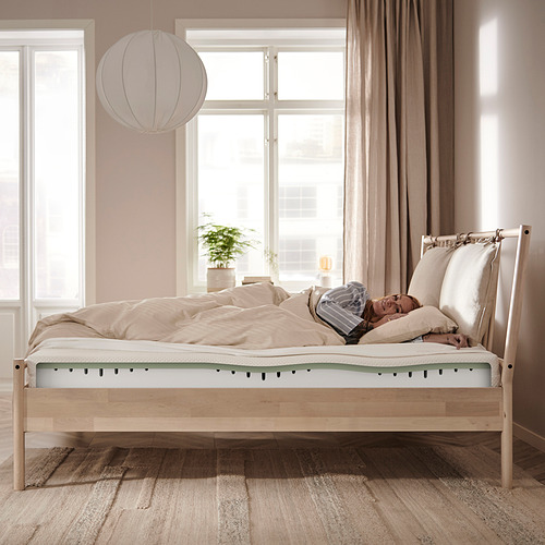 Un dormitorio sereno con una estructura de cama de madera clara con un firme colchón de espuma blanca y verde, una mesa auxiliar con una planta y una lámpara colgante.