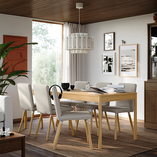 Una mesa de comedor de roble TONSTAD con seis sillas, colocada en el centro, moderna y espaciosa, iluminada por un gran ventanal, con auriculares y documentos.