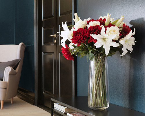 Jarrón de vidrio transparente con lirios blancos y flores rojas sobre mesa de madera, armario negro, silla gris. Decoración moderna de la habitación.