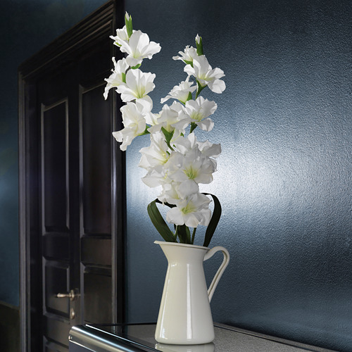 Flores artificiales blancas en jarrón, colocadas cerca de la puerta marrón oscuro en la pared azul.