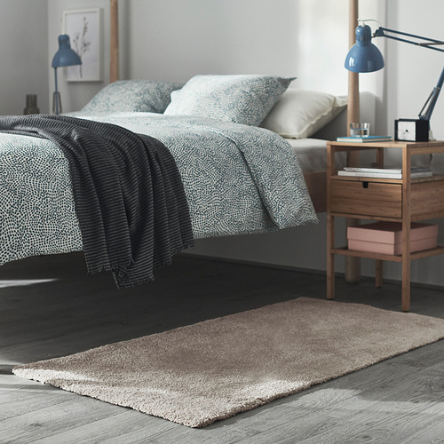 Dormitorio con cama, mesita de noche, lámpara y alfombra beige. Cama cuidadosamente hecha con almohadas. La mesita de noche tiene libros y una lámpara. La alfombra es suave y gruesa.