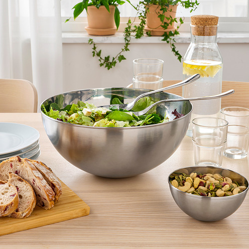 Gran cuenco de acero inoxidable con ensalada, pinzas, pan, frutos secos, vasos y jarra de agua sobre una mesa de madera.