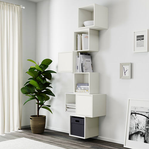 Las estanterías modulares blancas montadas en la pared organizan los libros y la decoración cerca de una ventana, con una planta alta y verde a su lado.