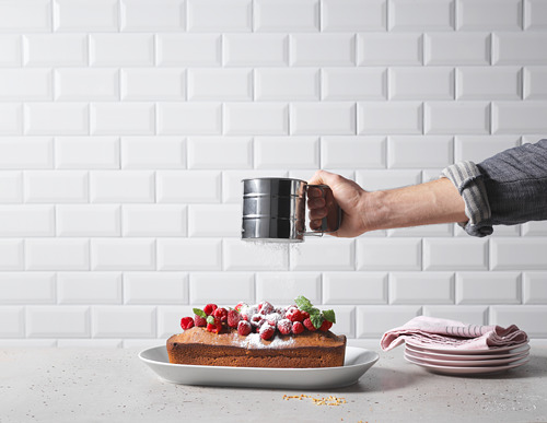 Una mano que sostiene un tamiz negro IDEALISK espolvorea azúcar en polvo sobre un pastel cubierto con bayas contra una pared de azulejos blancos.
