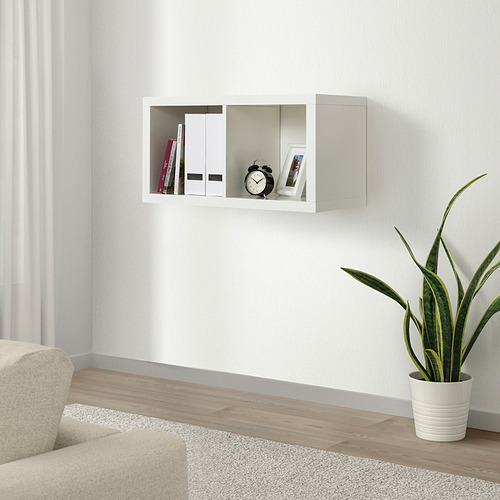 Los estantes de pared blancos KALLAX sostienen libros, reloj y fotos. Diseño minimalista de líneas limpias. Junto a una planta en maceta y una silla.