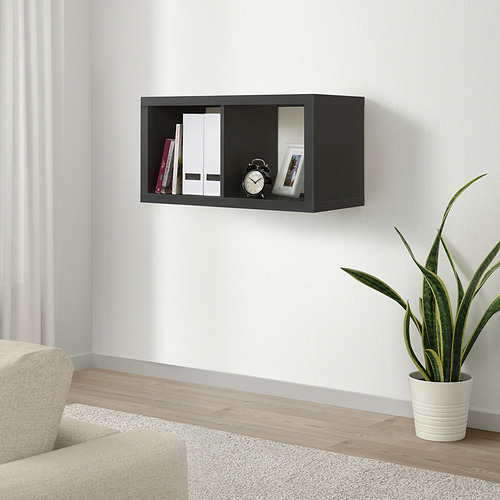 Un estante negro de pared KALLAX que contiene libros, un reloj y una foto enmarcada. Un sofá de color crema y una planta en maceta están debajo.