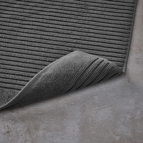 Alfombra de baño texturizada de color gris oscuro con esquina doblada sobre suelo de piedra gris.