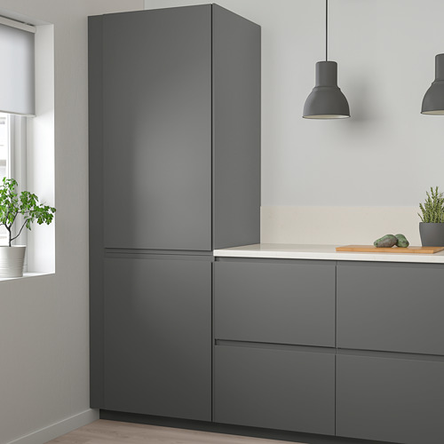 Cocina moderna con armarios VOXTORP de color gris oscuro, nevera grande a juego, manijas elegantes y plantas altas junto a la ventana.