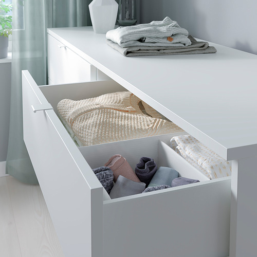 Cómoda blanca con ropa doblada. Textiles gris claro y beige cuidadosamente organizados. Diseño minimalista para dormitorio o salón.