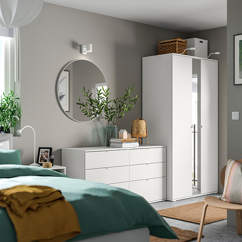 Dormitorio moderno con ropa de cama verde azulado, muebles blancos, espejo redondo y planta verde.