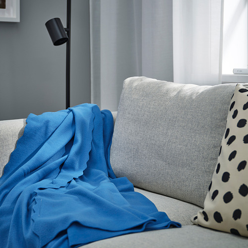 Una acogedora manta de forro polar azul DAGGKÅPOR, que cubre predominantemente un sofá gris claro, con almohadas de lunares negros y una lámpara cerca.