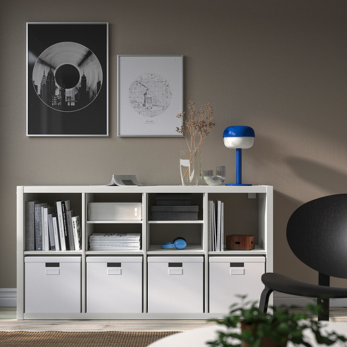 Estantería KALLAX blanca con cajones, libros, lámpara y pósters, silla negra.