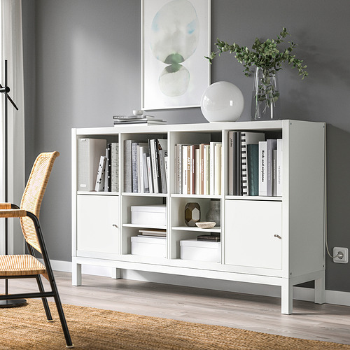 Estantería KALLAX con libros y decoración contra la pared gris, silla de mimbre a la izquierda, se eleva para facilitar la limpieza.