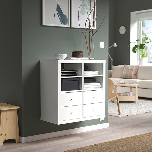 Un mueble KALLAX blanco contra una pared verde con estantes y cajones, que exhiben diversos artículos. Un ambiente acogedor en la sala de estar, reloj y plantas notables.