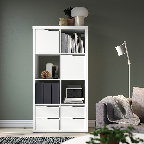 Estantería blanca KALLAX con libros, jarrones y plantas contra una pared verde junto a una silla y una lámpara grises.
