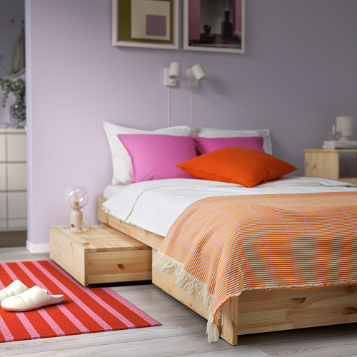 Almohadas naranjas y rosas sobre cama de madera, alfombra roja, zapatillas, lámpara lateral, paredes moradas con arte.