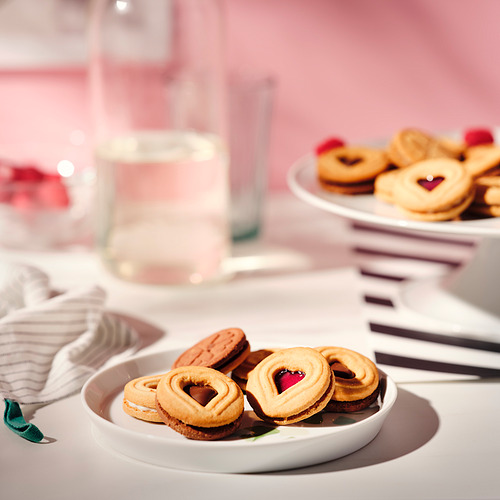 Dos platos de galletas de frambuesa y chocolate en forma de corazón, colocadas sobre un mantel rosa con vasos y servilletas.