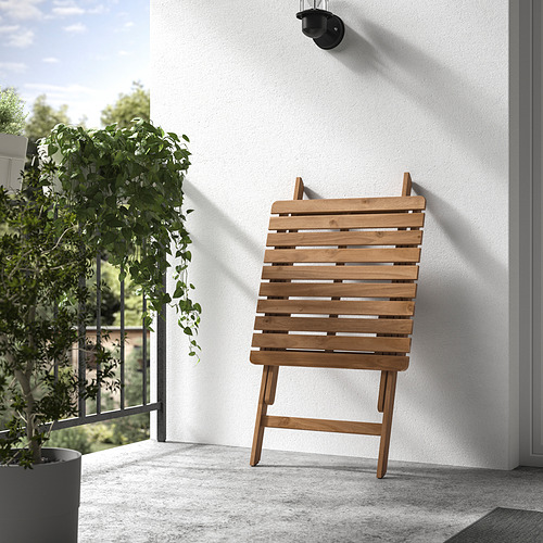Silla plegable de madera contra una pared blanca en un balcón, con una planta colgante al lado.