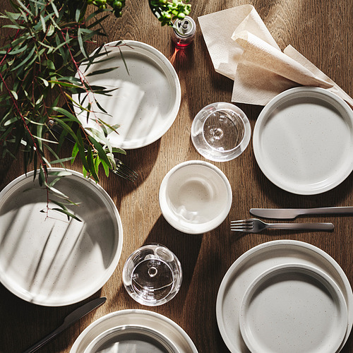 Plato de gres blanco colocado sobre mesa de madera con vasos, cubiertos y plantas. Los platos son más grandes que los vasos y las servilletas.