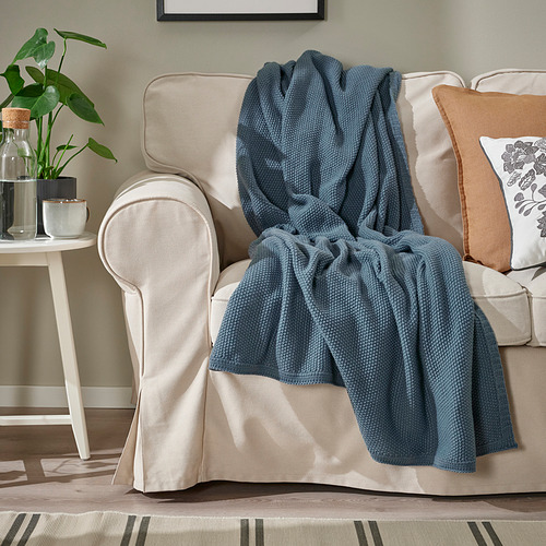 Una acogedora sala de estar con un sofá beige, decorado con una suave manta y almohadas de color gris azulado, y una mesa auxiliar con una planta.