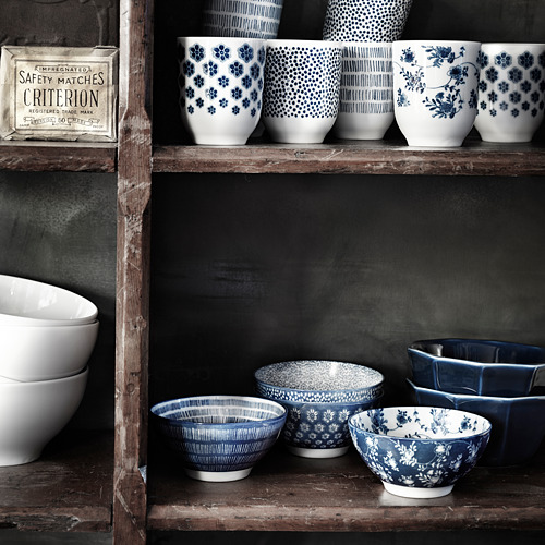 Colección de tazas y cuencos de cerámica azul y blanca con diseños florales y geométricos, dispuestos sobre estanterías de madera rústica.