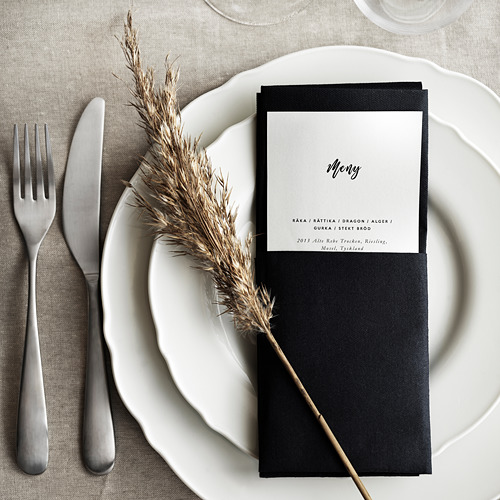 Tarjeta de menú negra en plato con volantes blancos, tenedor y cuchillo plateados, tallo de planta seca, mantel gris.