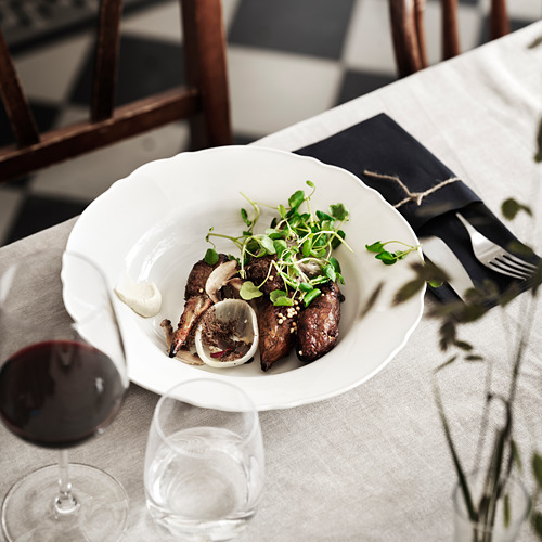 Un plato blanco con comida a la parrilla y verduras en una mesa con vino y cubiertos.