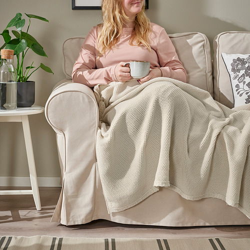 Mujer sentada en una silla beige, manta sobre las piernas, sosteniendo una taza de café.