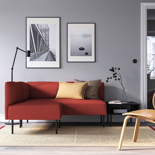 Sala de estar moderna con un gran sofá modular LILLEHEM rojo, mesa auxiliar negra, silla de madera y arte de pared. Las paredes grises resaltan una configuración acogedora.