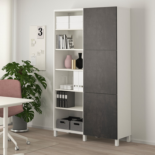 Mueble de almacenamiento BESTÅ blanco y gris con compartimentos abiertos y cerrados, cerca de la planta y la silla.