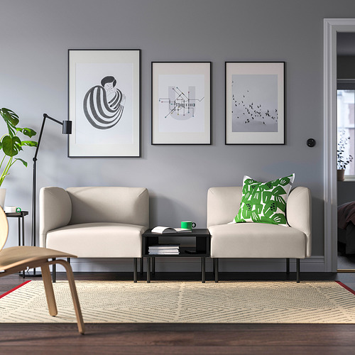 Sala de estar moderna con sofás neutros, almohadas verdes, mesa de centro negra, arte abstracto, paredes grises, suelo de madera, alfombra y lámpara alta con planta.