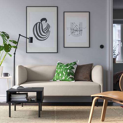 Fokus: Sofá modular beige LILLEHEM, pared gris, almohadas verdes, mesa de centro negra, silla de madera, estampados de arte moderno.