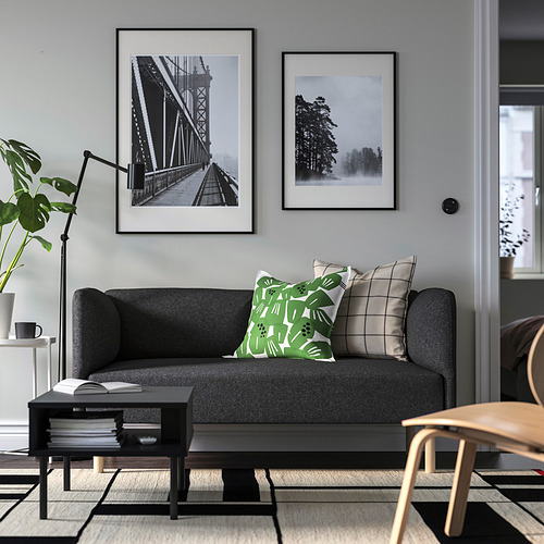 Sala de estar moderna con sofá gris, almohadas verdes/neutras, fotos negras, mesa auxiliar con libros, silla y planta.