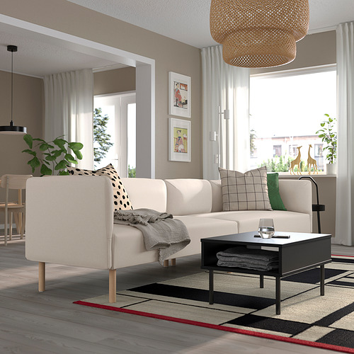 Moderno salón con sofá LILLEHEM, mesa de centro negra, alfombra, plantas y gran ventanal.