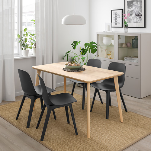 Pequeño comedor moderno con mesa rectangular de madera clara, cuatro sillas negras y lámpara de pie ligera. Gran ventanal con cortinas, armario y plantas.