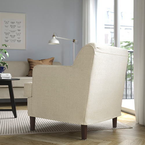 Sillón beige con patas de madera en acogedora habitación; lámparas, sofá y arte visibles.