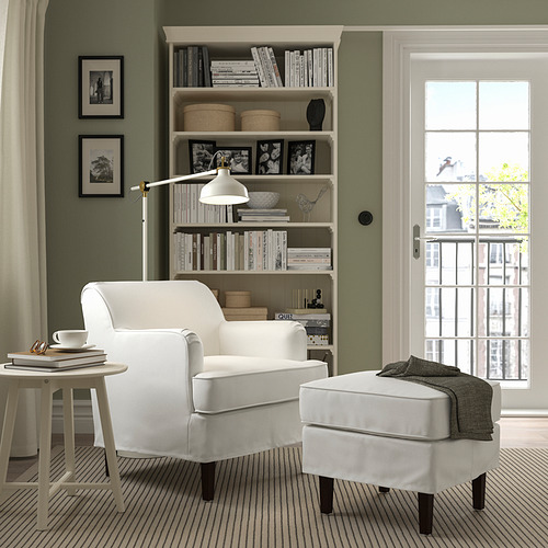 Acogedora sala de estar con sillón ROCKSJÖN, reposapiés y estante lleno de libros. Paredes verdes, luz natural y un reposapiés cubierto de suéter.