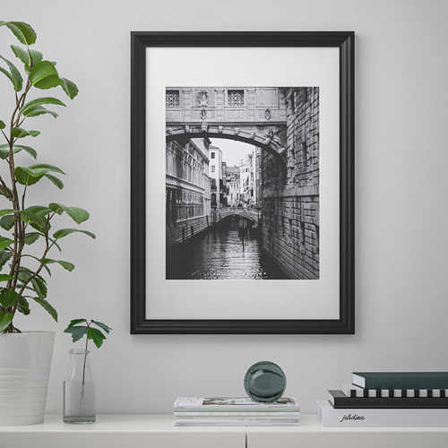 Un puente y un canal en blanco y negro con marco negro cuelga de una pared. En un estante de abajo, hay libros, una planta, un jarrón y un objeto redondo.