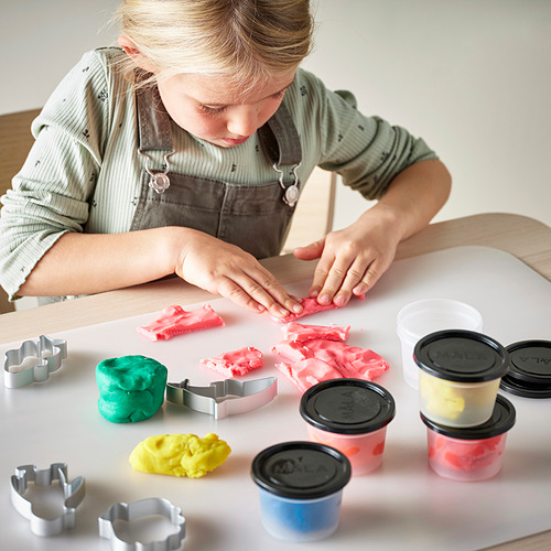 Un niño juega con masa de colores para modelar en una mesa, usando cortadores de galletas para dar formas. Varios recipientes con tapas contienen masa de diferentes colores.
