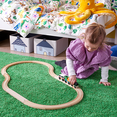 Un niño pequeño juega con un tren magnético de madera colocado sobre una alfombra verde debajo de una cama con un pulpo amarillo de peluche.