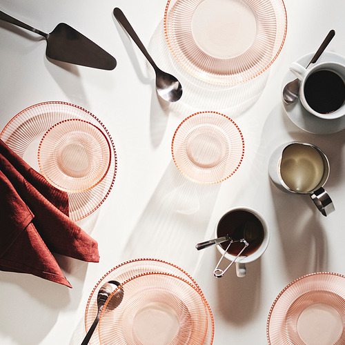 Elegante montaje de mesa con cristalería y utensilios rosas. Platos, tazas, servilletas y servidor de pasteles redondos con textura.