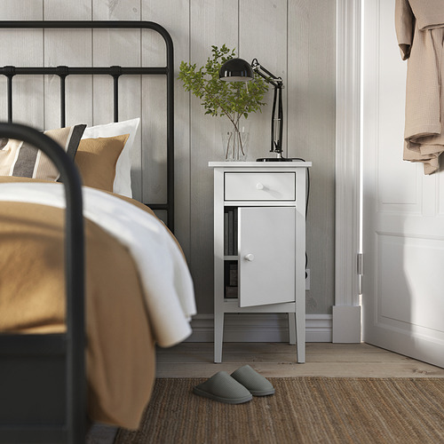 Dormitorio con mesita de noche GULLABERG blanca, lámpara negra, zapatillas verdes y cama con sábanas doradas y blancas.