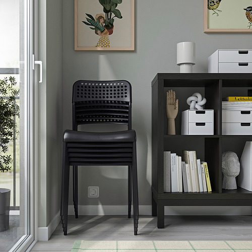 Silla arenquera apilable negra cerca de estantería negra con libros, decoración y arte en la pared gris.