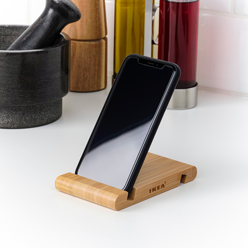 Un teléfono inteligente negro descansa sobre un soporte de madera con grabado de Ikea, colocado en posición vertical sobre una superficie blanca, rodeado de elementos decorativos.