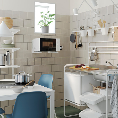 Pequeña cocina con microondas blanco, silla azul, fregadero de acero inoxidable, utensilios de madera, planta verde y pared de azulejos.