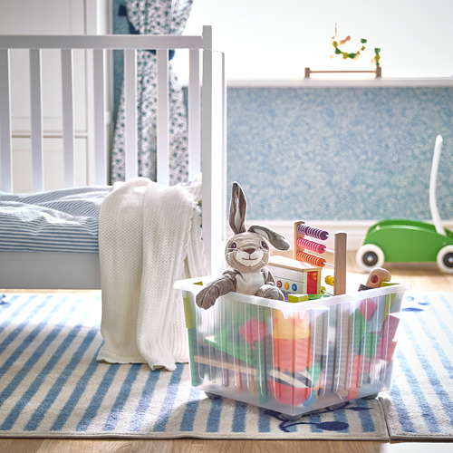 Habitación para niños con caja de juguetes VESSLA sobre alfombra azul al lado de la cuna.