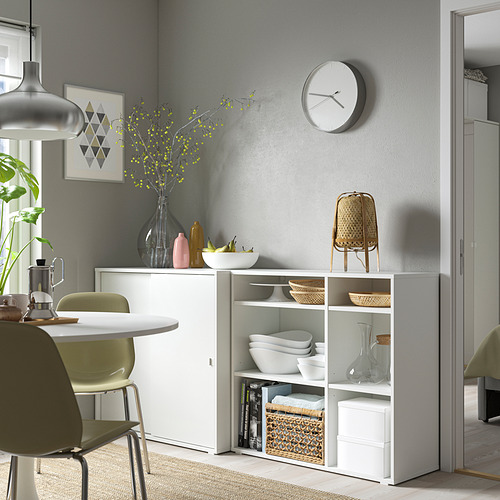 Moderno comedor con mueble de almacenamiento VIHALS blanco. Estantes con cuencos, jarrones y decoración. Sillas verdes, pared gris y reloj encima.