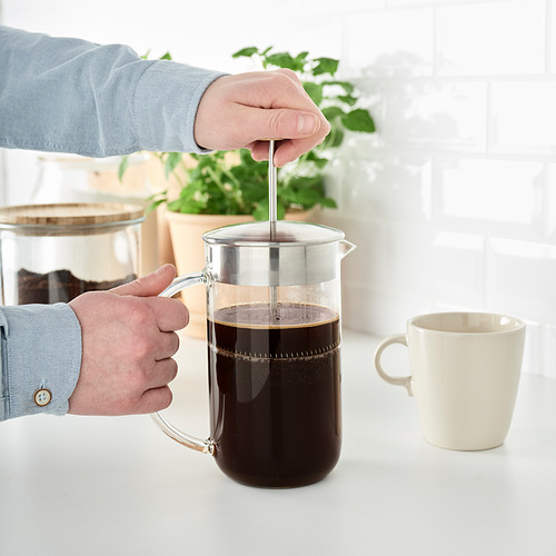 Una persona está presionando un émbolo en una cafetera de vidrio, extrayendo café en una jarra. Una taza blanca está al lado del fabricante.