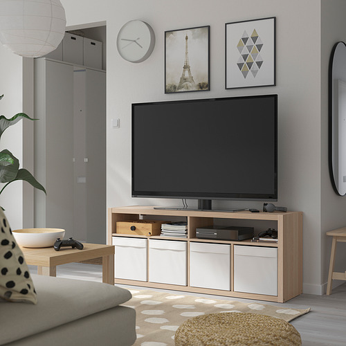 Banco de tv KALLAX en blanco y roble contra la pared, con cuadros enmarcados, reloj, sofá gris, alfombra y espejo redondo.